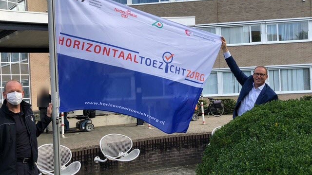 Bestuurder Ton Hazekamp hijst met hulp van beveiliger Heico Harmsen de vlag Horizontaal Toezicht in de mast van het Slingeland Ziekenhuis.